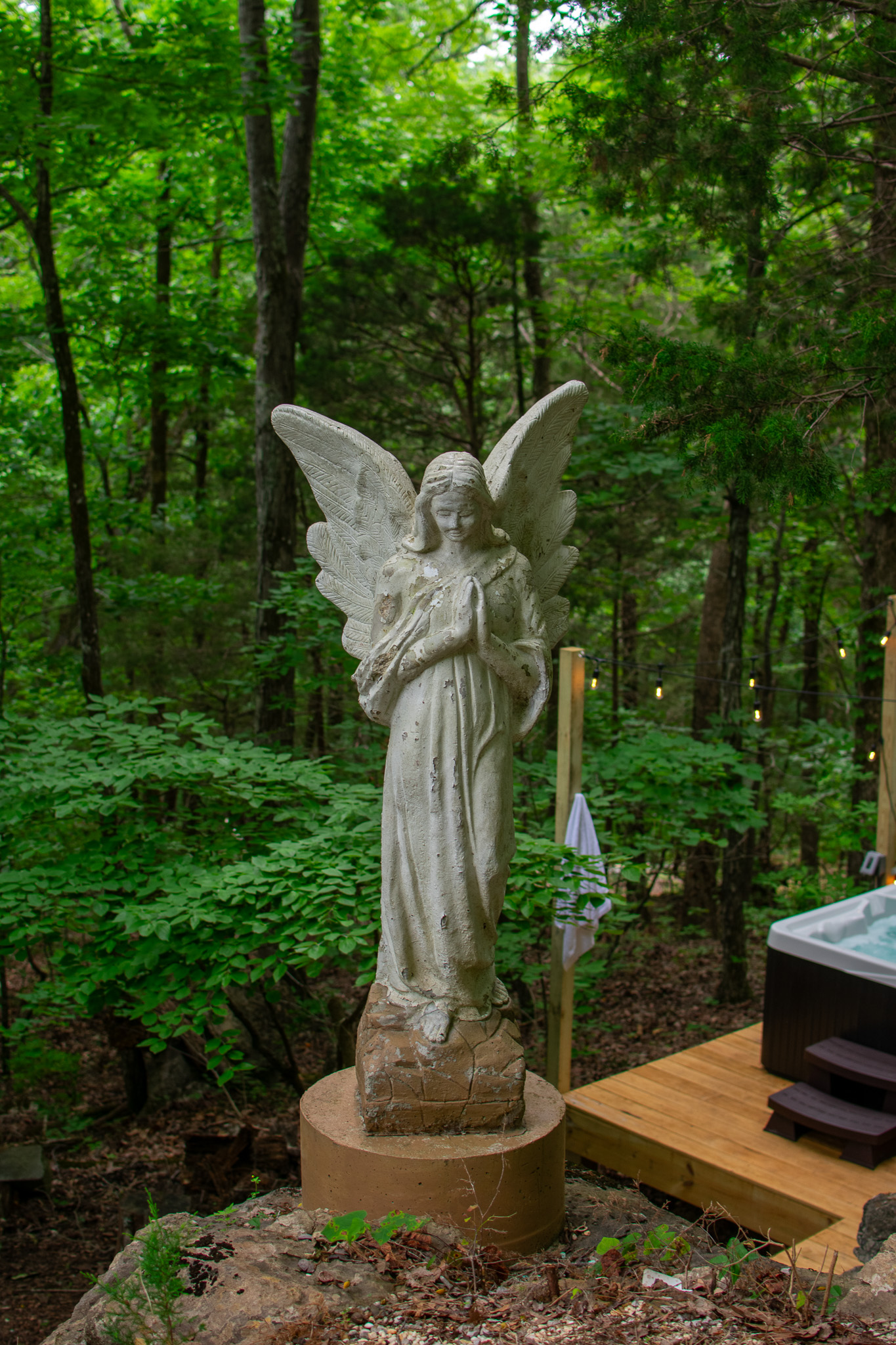 Weathered angel statue with folded hands standing on a round base in a wooded outdoor area.