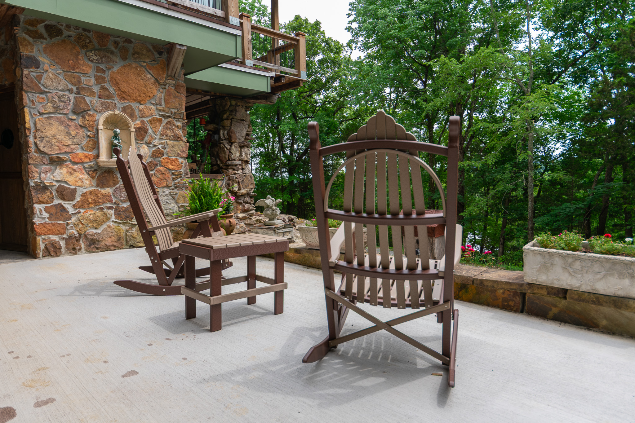 Two wooden rocking chairs and a small table on a concrete patio next to a stone house with trees beyond.