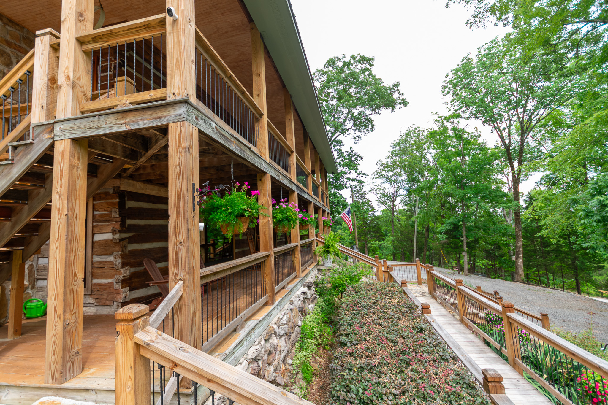 Wooden cabin porch with hanging flower baskets and an American flag by a forested pathway.