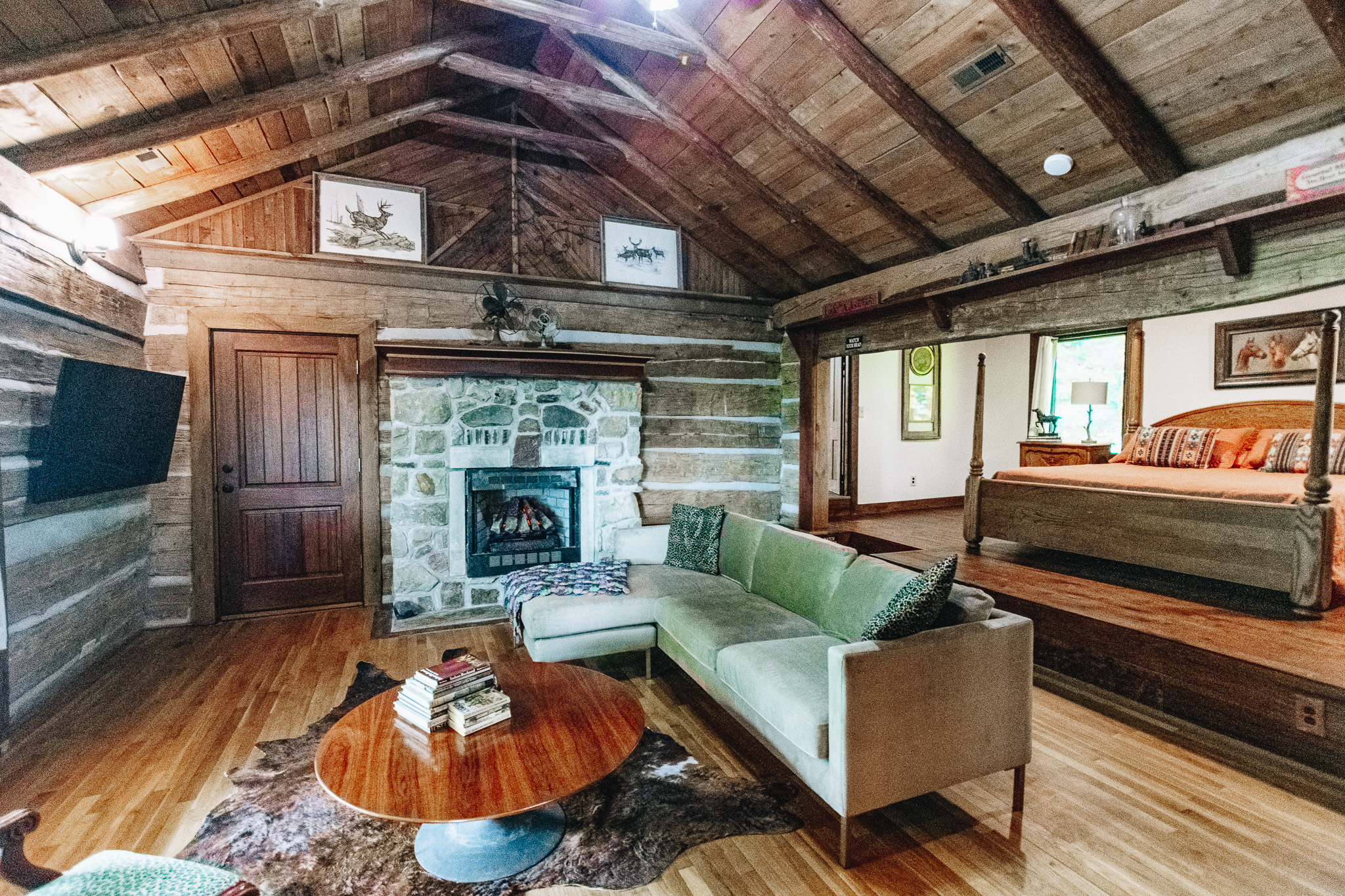 Cozy living room with wooden walls, green sectional sofa, stone fireplace, and adjoining bedroom with wooden bed.