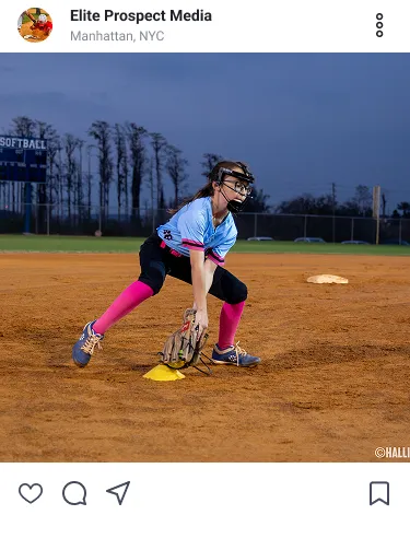 Female softball player in pink socks and blue jersey fielding a ground ball on a dirt softball field at dusk.