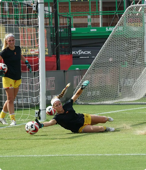 Female soccer goalie diving to catch a ball while teammate watches near the goalpost on grass field.