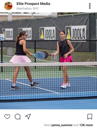 Two girls on a pickleball court tapping paddles mid-game, wearing athletic skirts and sneakers.