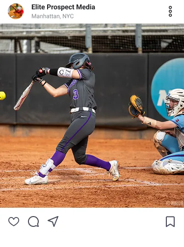 Female baseball player in a black and purple uniform swings bat at a yellow ball while catcher in blue crouches.