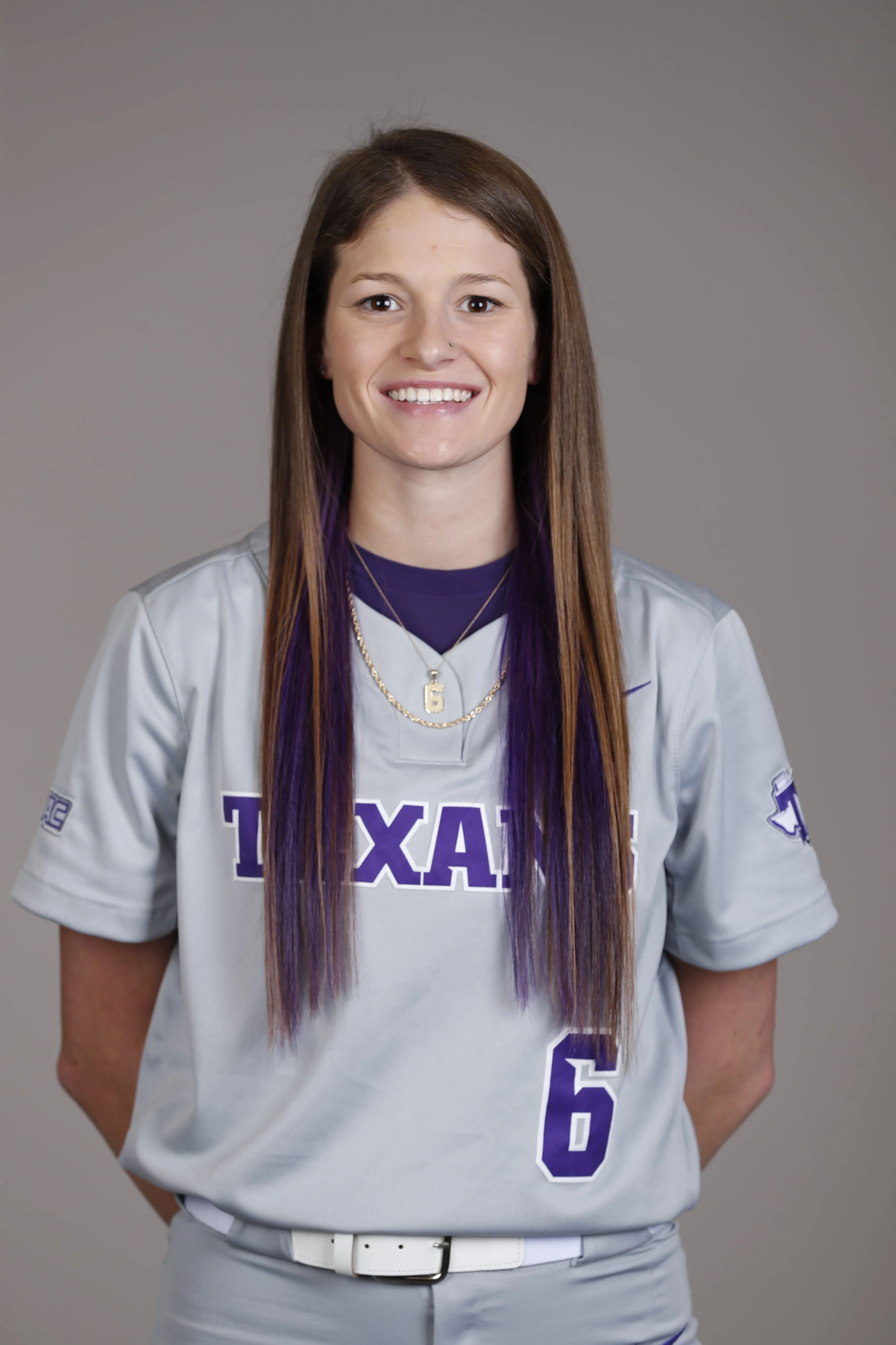 Smiling softball player in gray Texas jersey with number 6 and purple highlights in long hair.