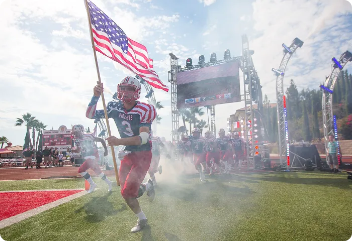 Football player holding US flag runs onto field with teammates behind through smoke and lights.