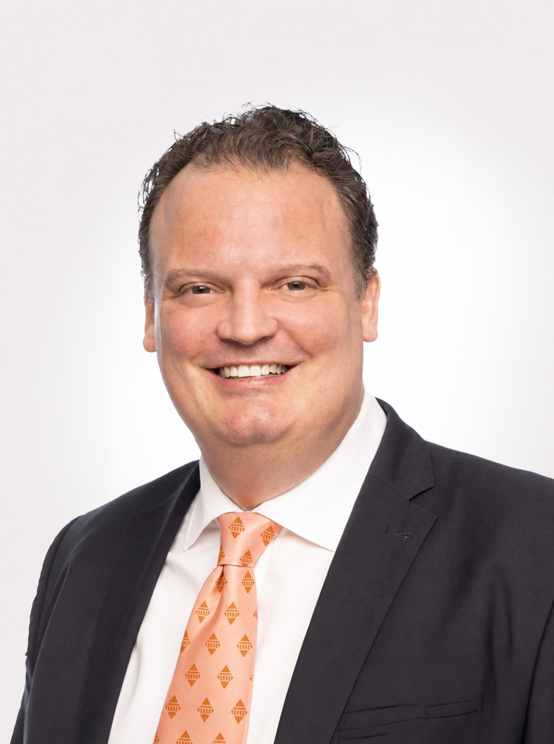 Smiling Dr. Roeder in a black suit, white shirt, and orange patterned tie against a white background.