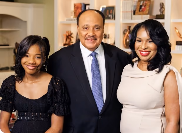 Three people, a young girl, a man in a suit with a blue tie, and a woman in a cream dress, smiling indoors.
