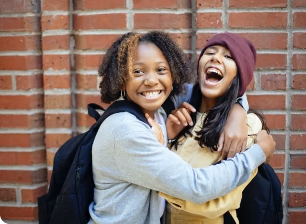 Two smiling young women with backpacks hugging joyfully in front of a brick wall.