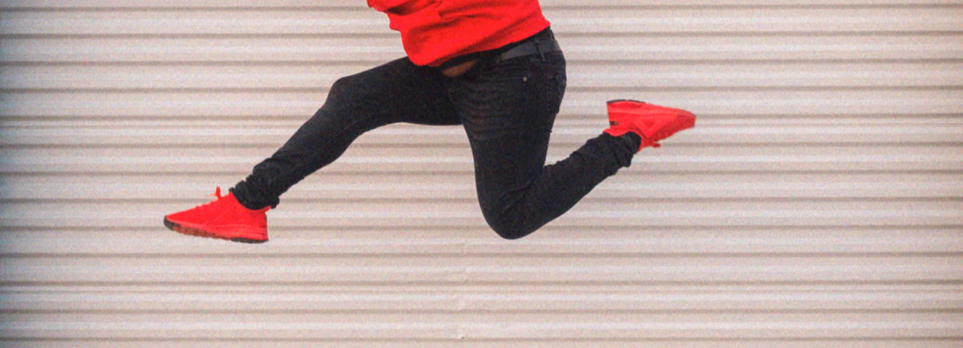 Person wearing red shoes and black pants jumping against a light horizontal-striped background.