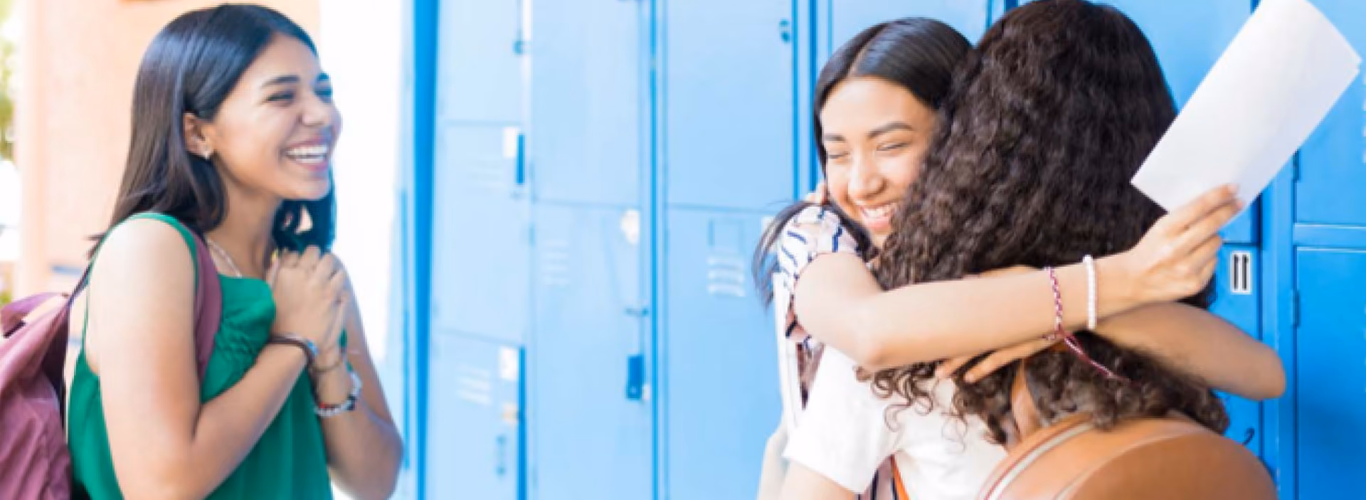 Two girls happily hugging near school lockers while another girl smiles at them.