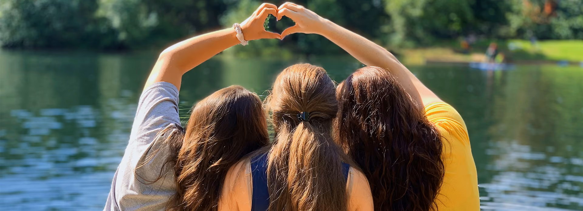 Three women sitting by a lake, making a heart shape with their raised hands.