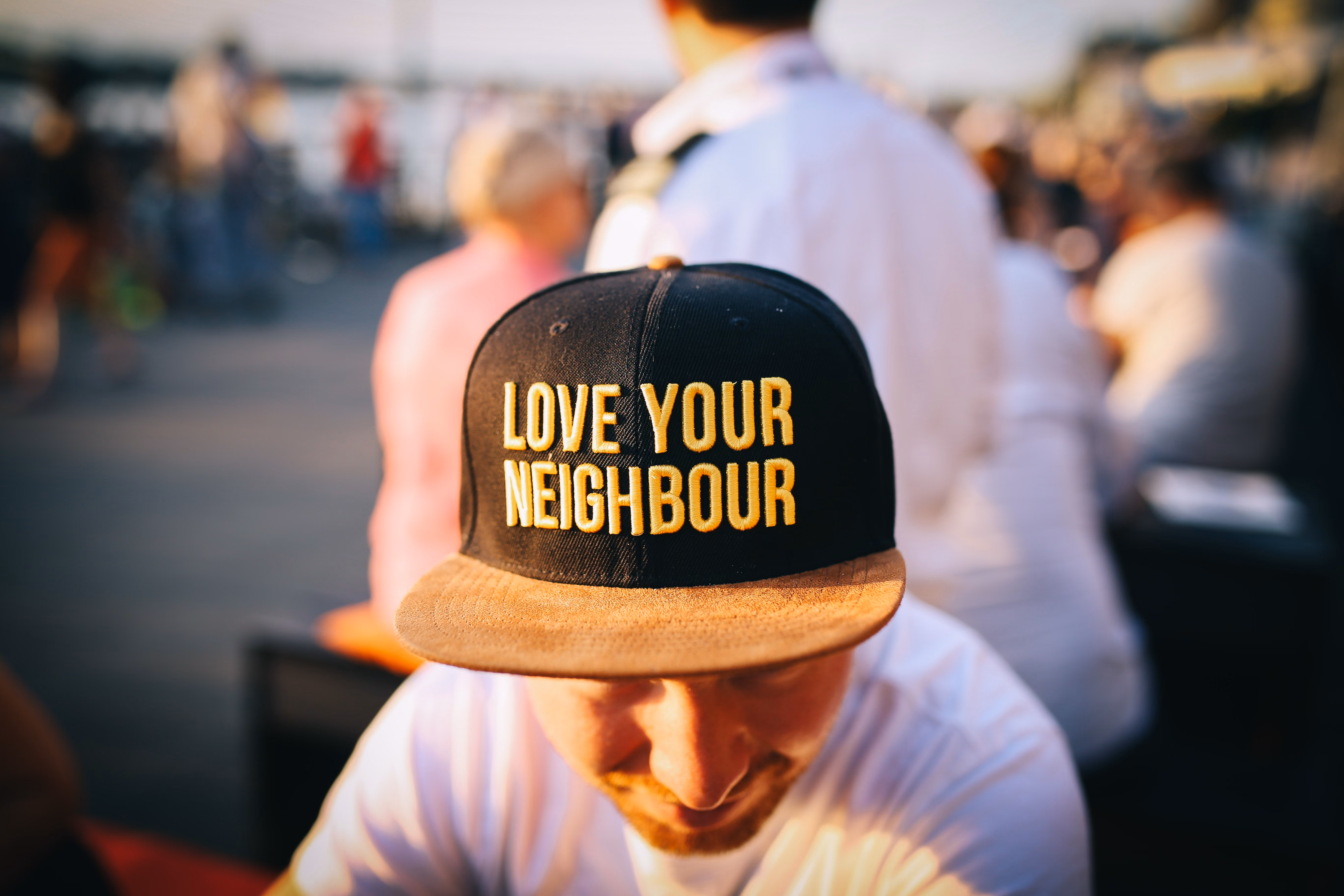 Man wearing black and brown cap with yellow text 'LOVE YOUR NEIGHBOUR' in an outdoor setting.