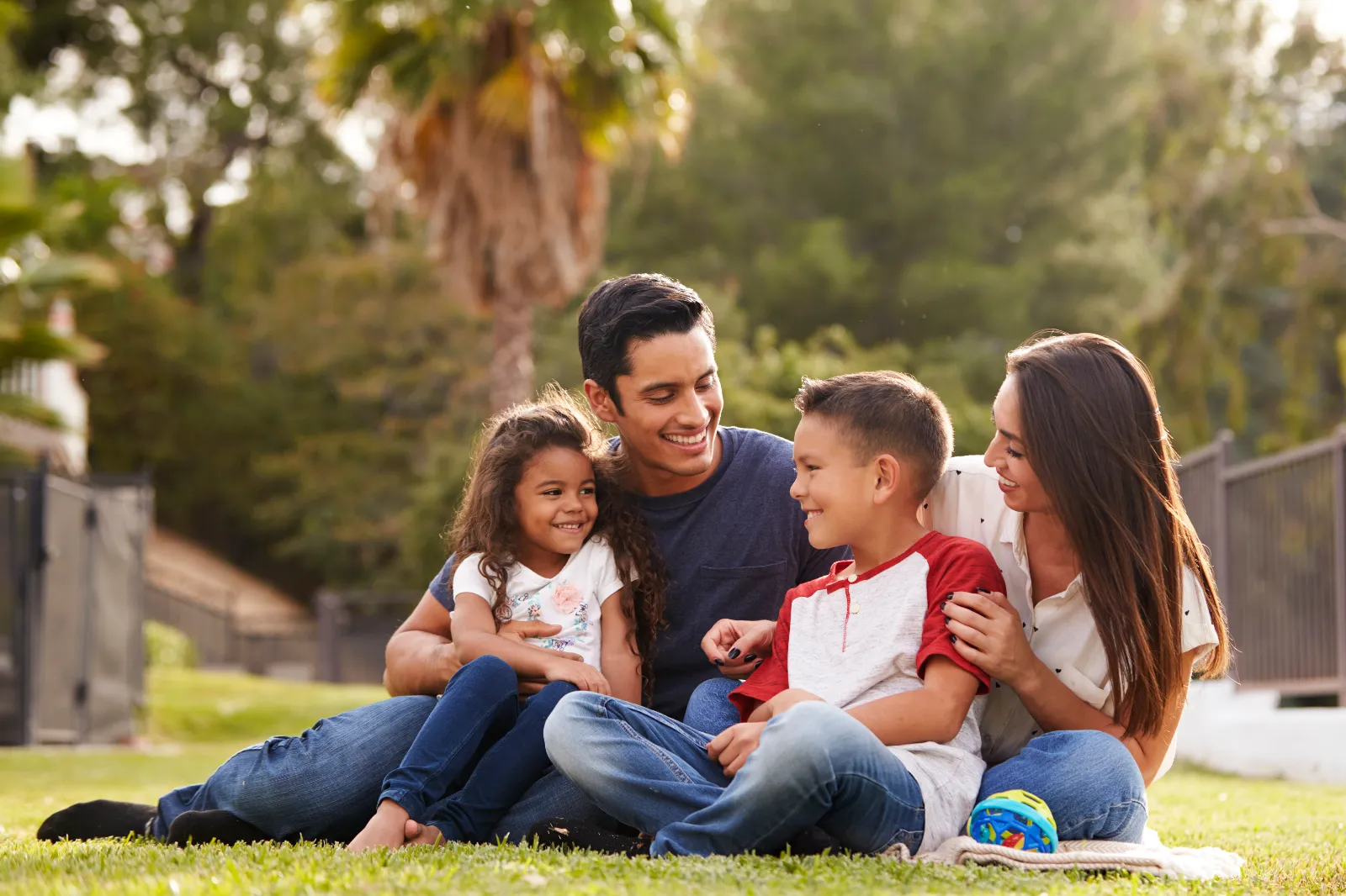 A father and mother sitting on the grass outdoors with their two young children, all smiling.