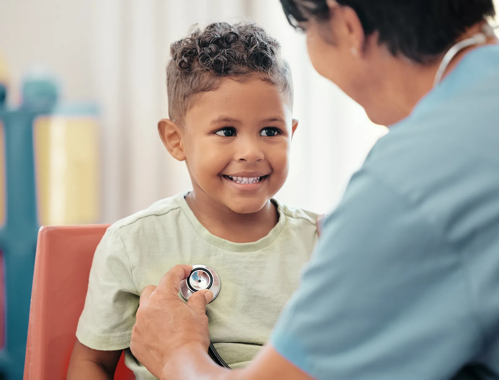 A smiling young boy having his heart listened to with a stethoscope by a healthcare provider.