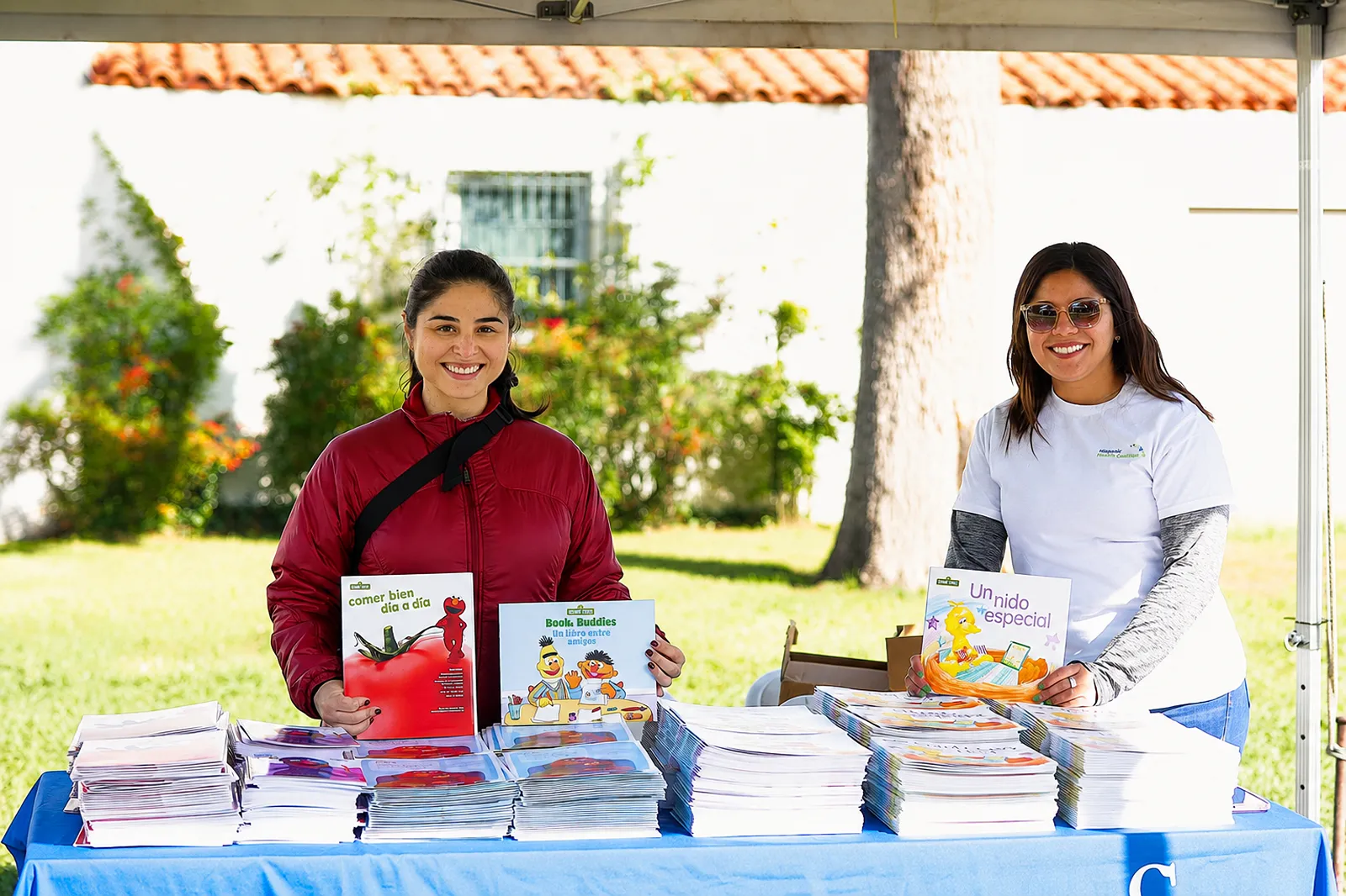 Two staff members standing behind a table stacked with Spanish-language educational booklets at an outdoor community event tent.