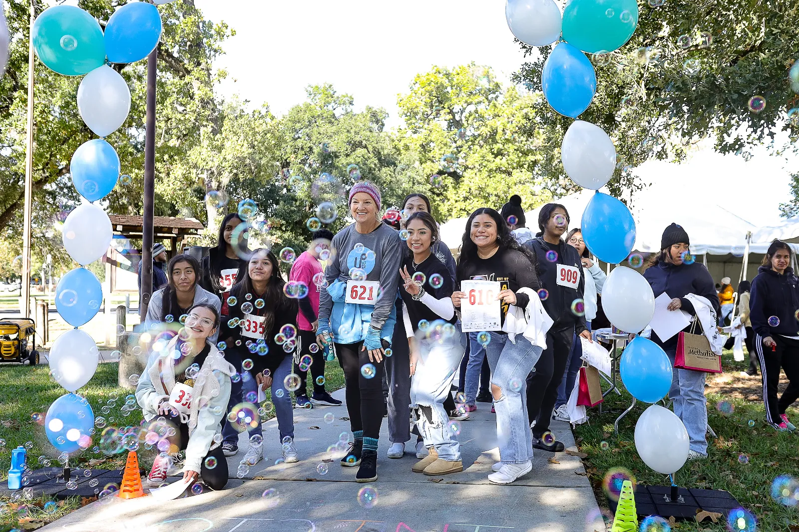 A group of participants wearing race bibs posing under a blue and white balloon arch at an outdoor community walk or run event, with bubbles floating around them.