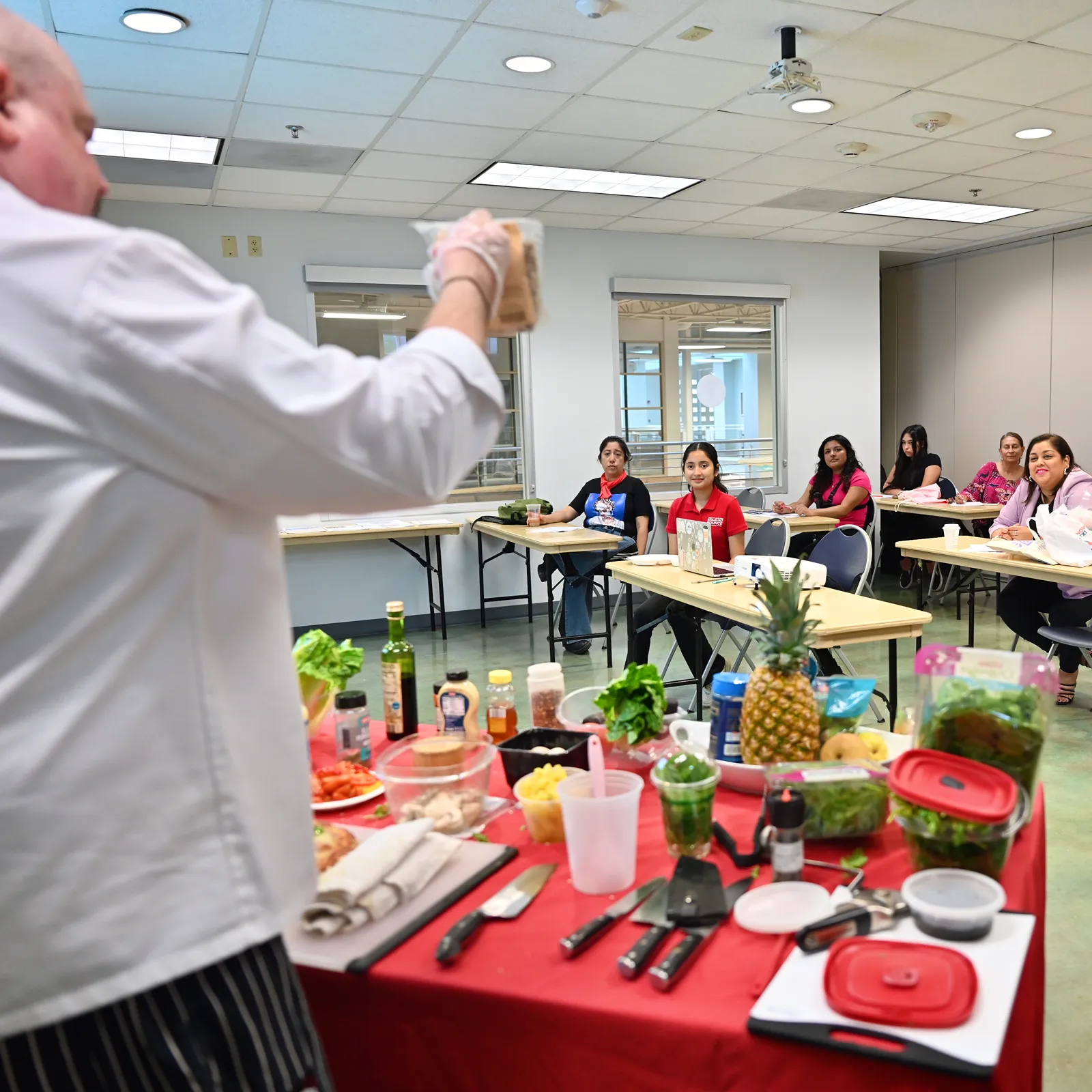A chef leading a healthy cooking demonstration in front of a seated audience, with fresh produce and utensils laid out on the table.