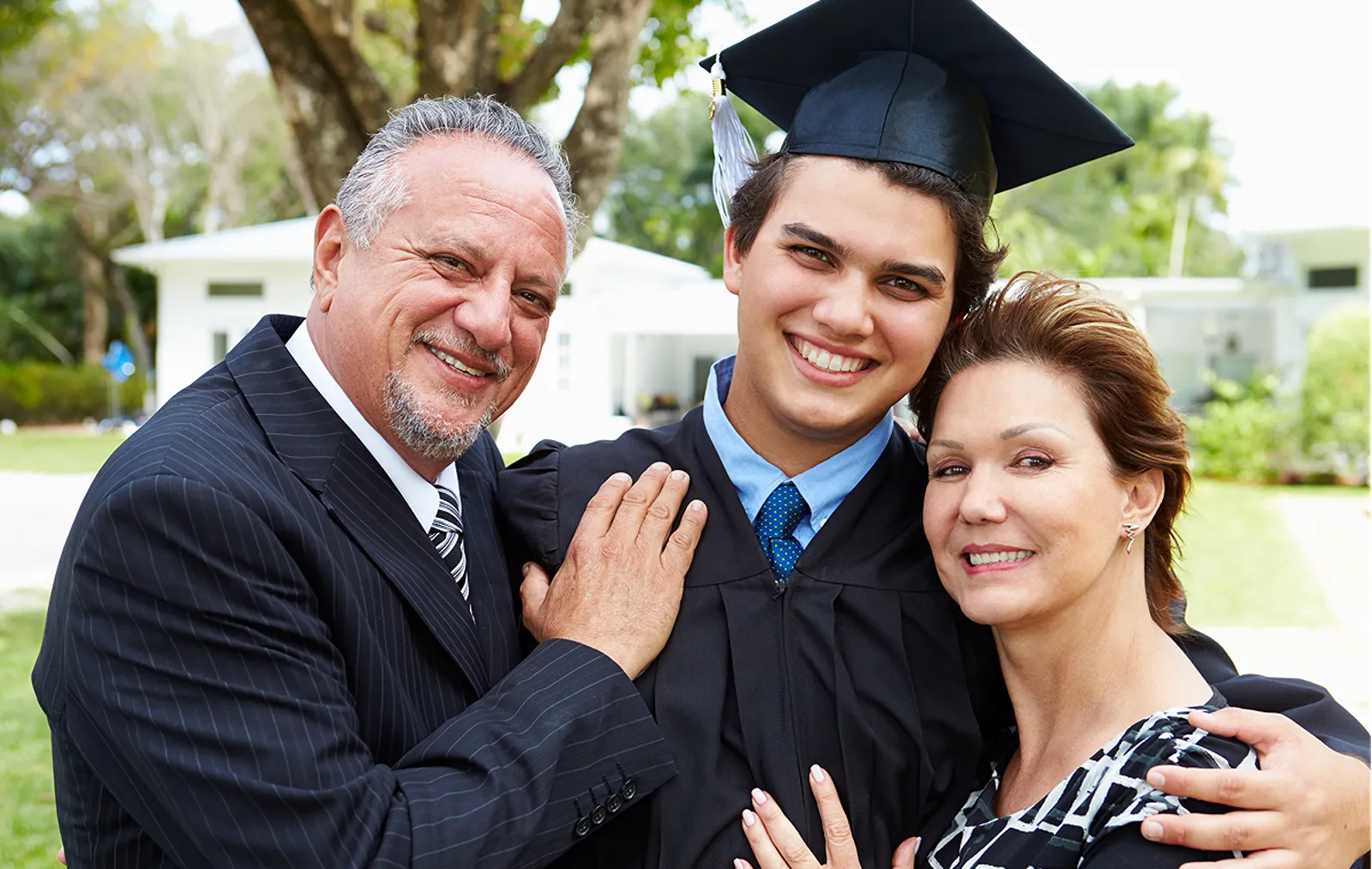A young man in a graduation cap and gown posing outdoors with his proud parents.