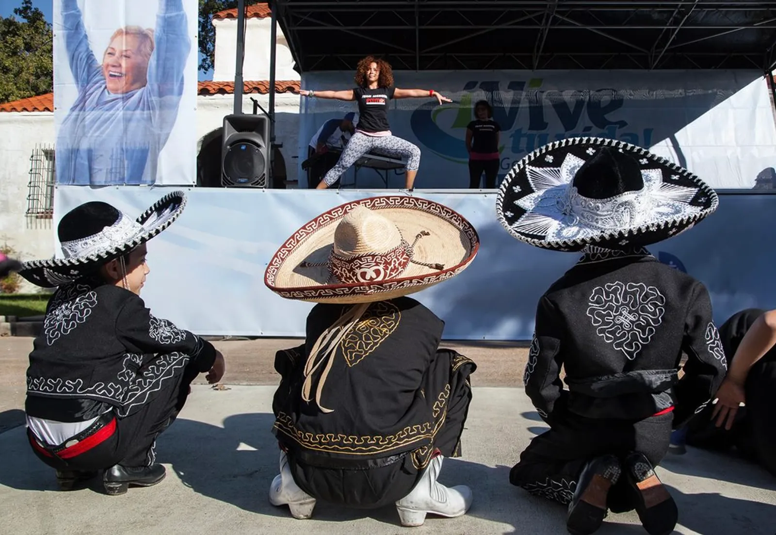 Mariachi performers in traditional charro sombreros watching a fitness instructor lead an exercise routine on stage at a 'Vive tu vida' community event.