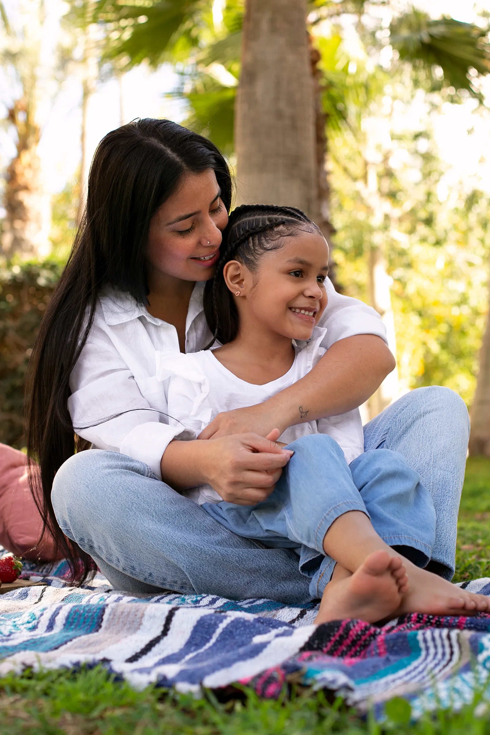 A mother embracing her young daughter while sitting together on a blanket outdoors in a park.