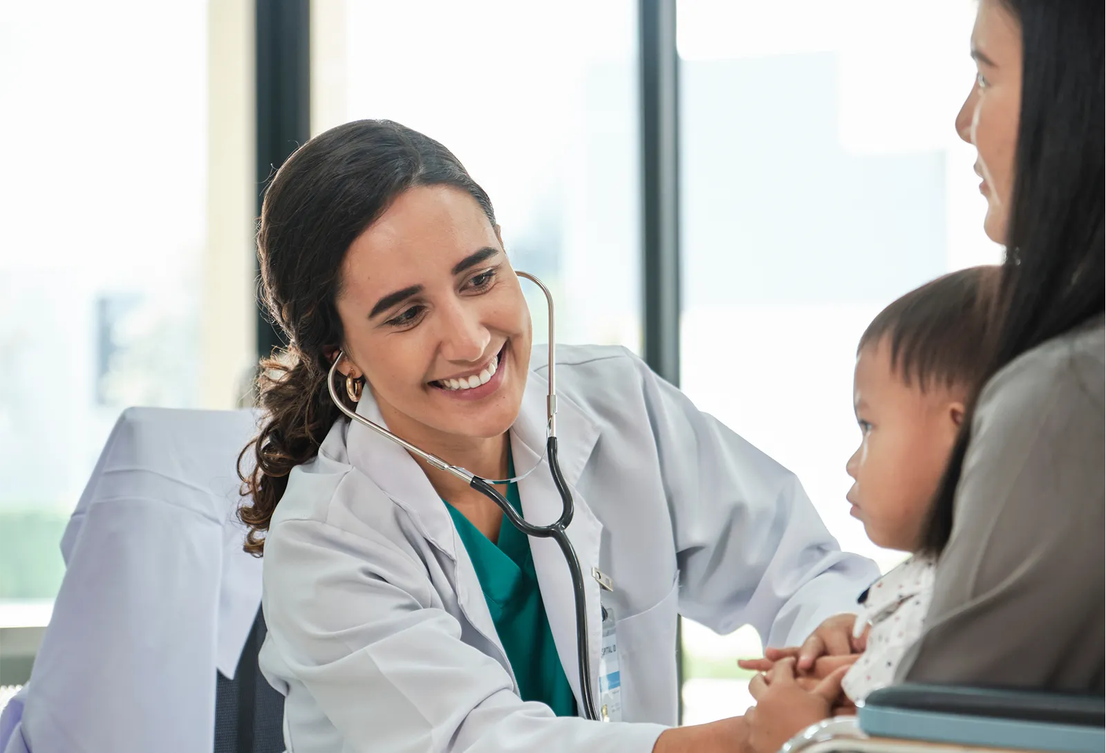 A smiling doctor with a stethoscope leaning in to greet a toddler seated on a parent's lap during a checkup.