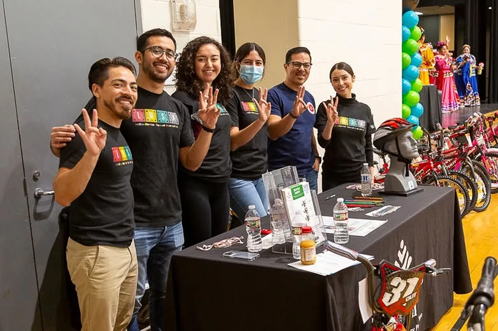 Six volunteers posing at an event table with bicycles and balloon decorations in the background.