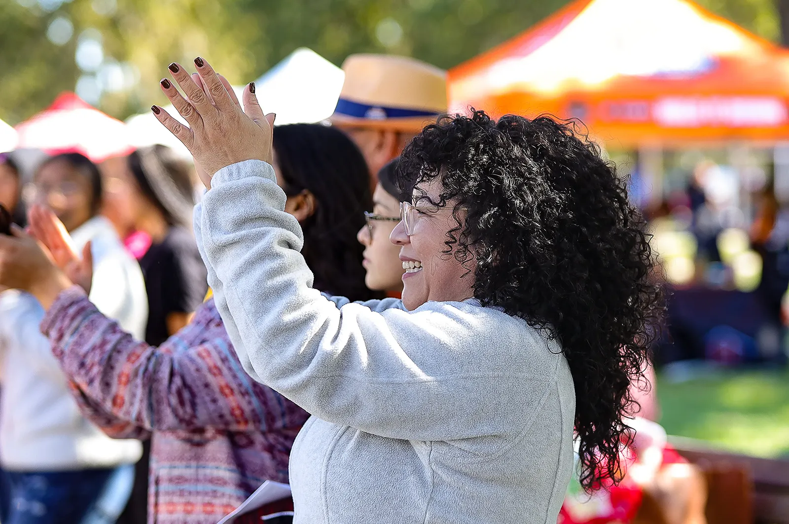 A woman with curly hair raising her hand while participating in an outdoor community event with tents in the background.