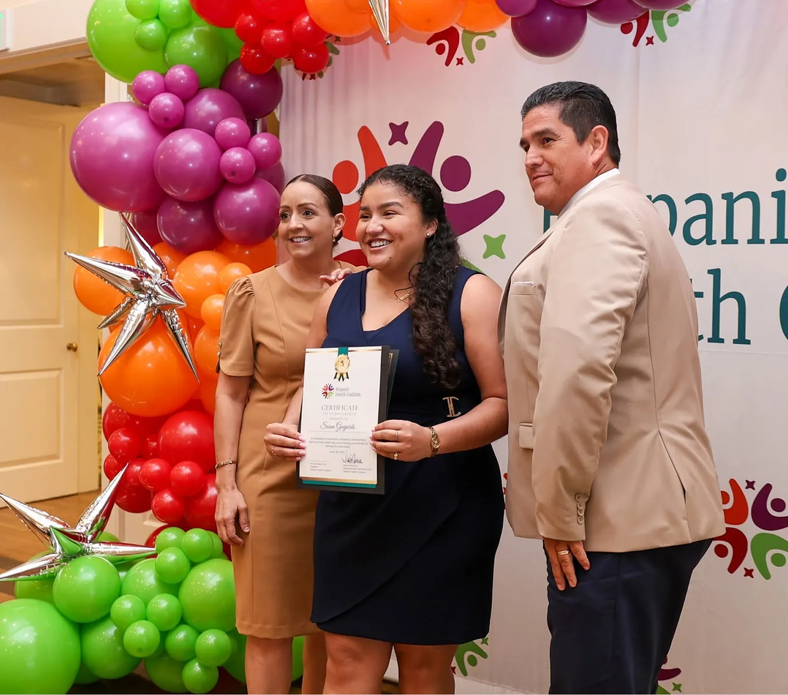 A young woman holding a certificate, flanked by two staff members in front of a Hispanic Health Coalition backdrop and balloon arch.