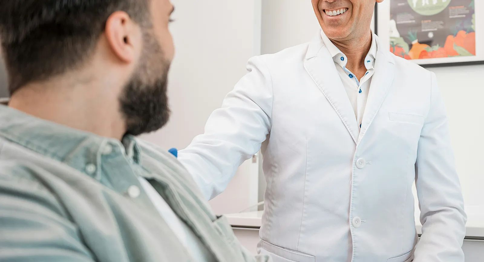 A smiling doctor in a white coat placing a reassuring hand on a patient's shoulder during a consultation.