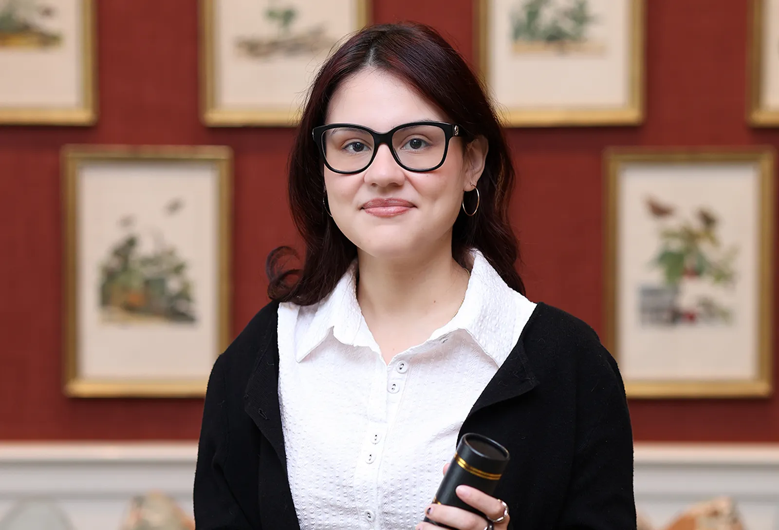 Portrait of a woman holding a microphone, standing in front of a wall of framed artwork.