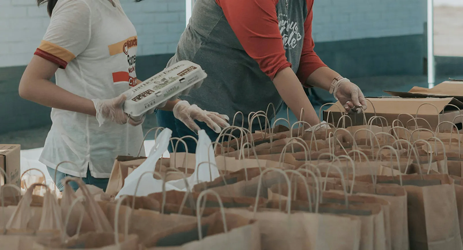 Volunteers wearing gloves packing rows of brown paper bags for a community food distribution.