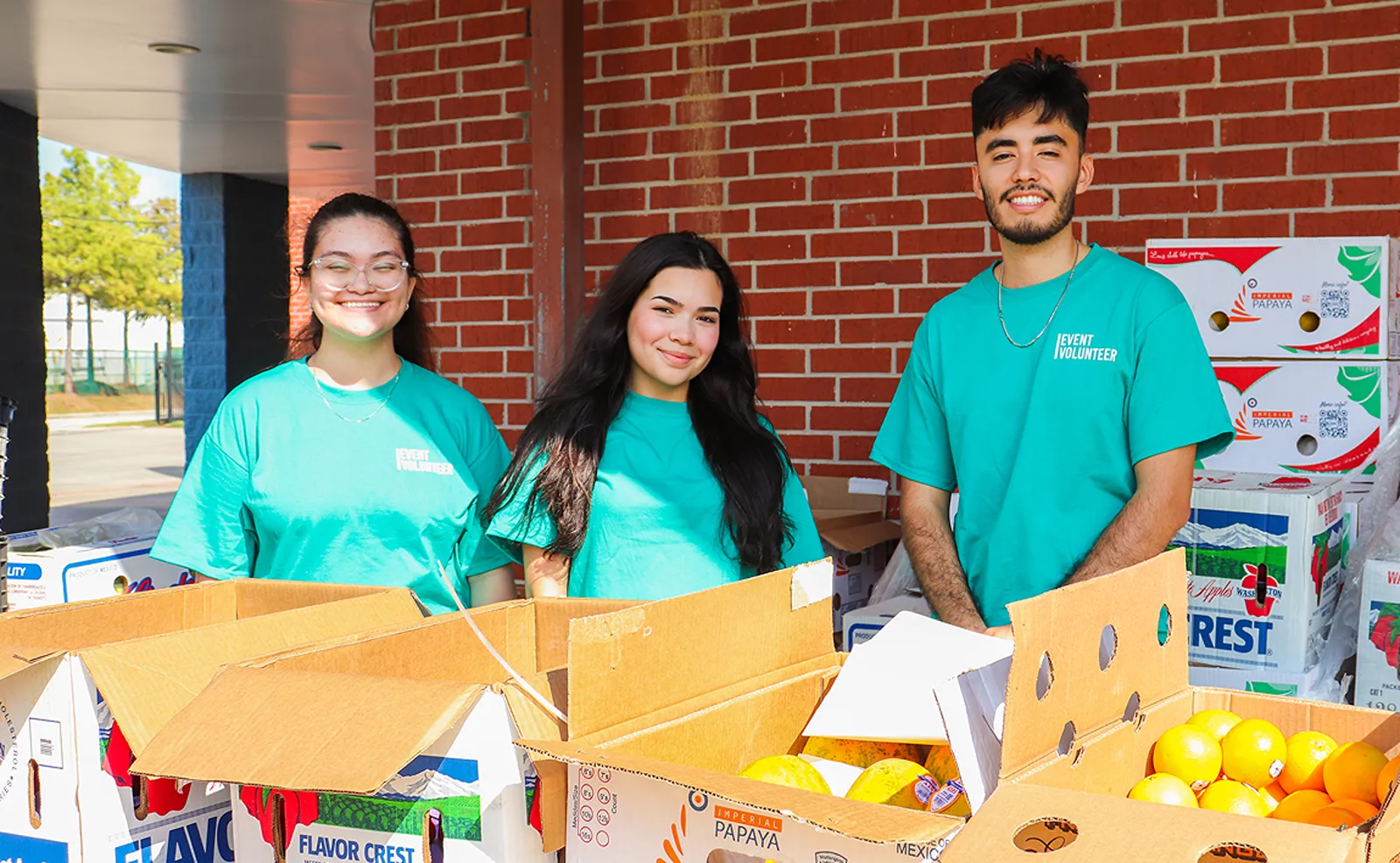 Three volunteers in teal shirts standing behind boxes of produce at a food distribution event.