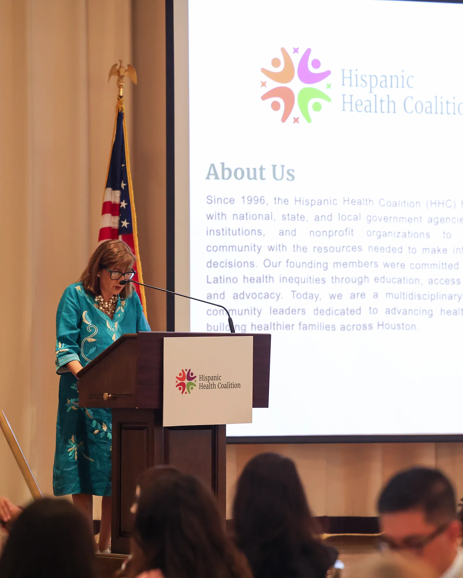 A woman speaking at a podium in front of a screen showing the Hispanic Health Coalition 'About Us' slide.