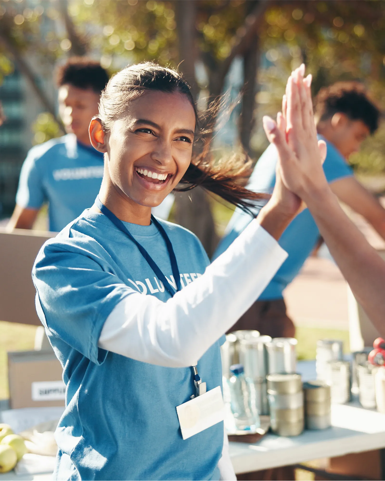 A smiling volunteer in a blue T-shirt high-fiving someone at an outdoor food distribution table.