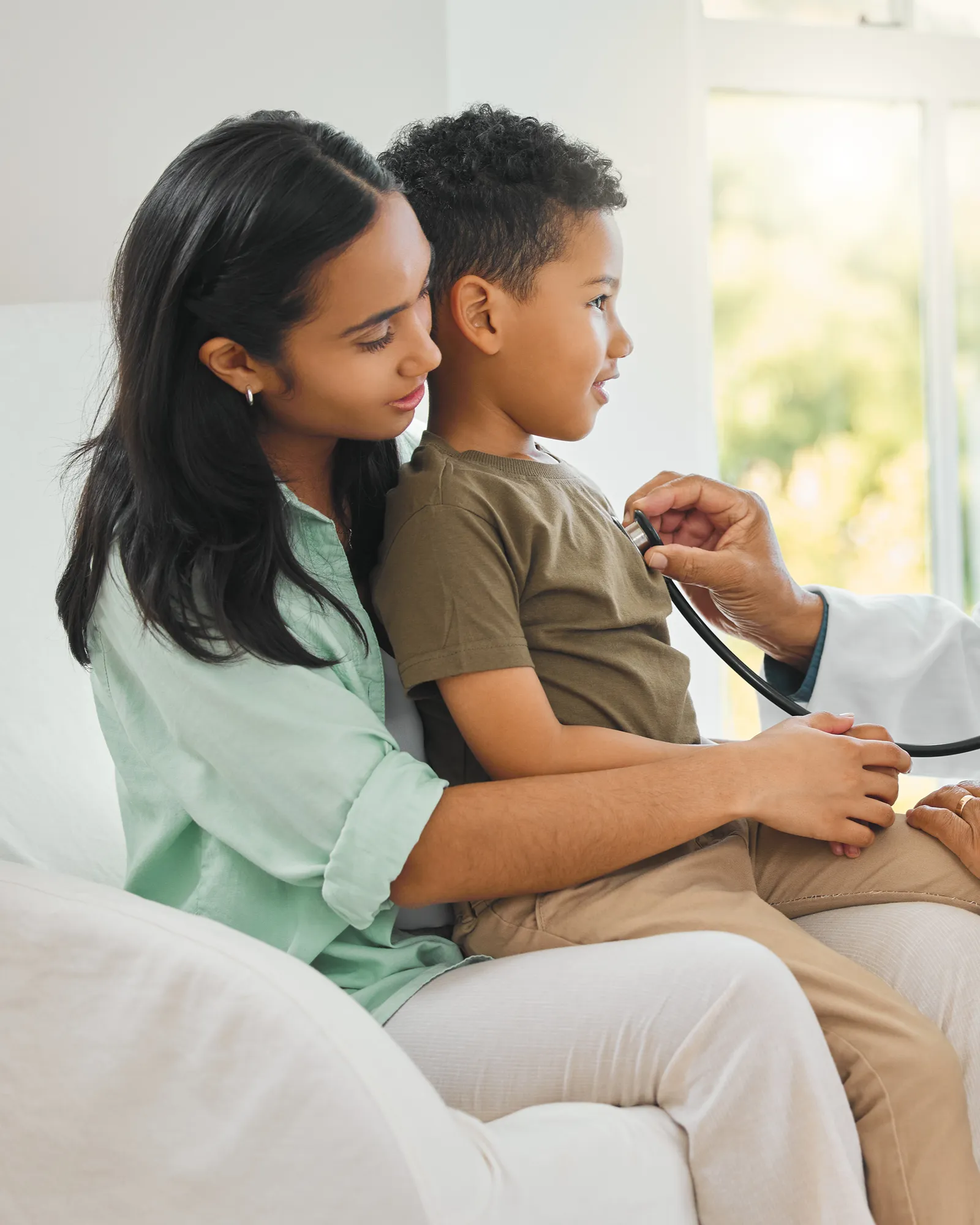 A doctor listening to a young boy's back with a stethoscope while he sits on his mother's lap.