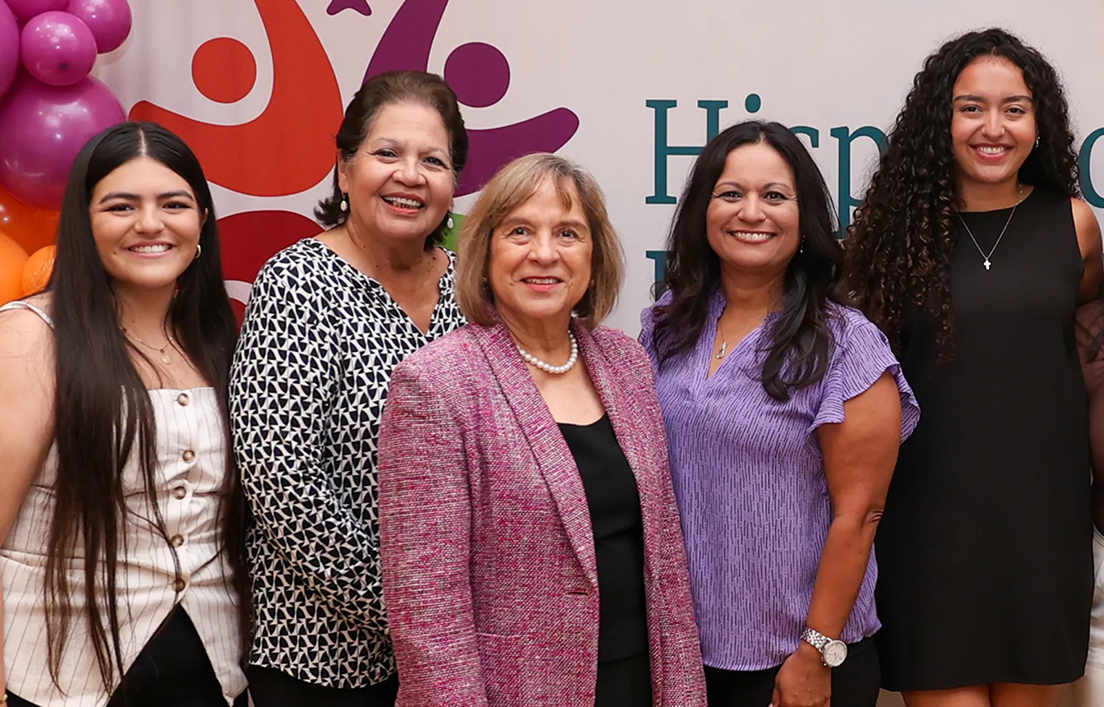 Five women posing together in front of a Hispanic Health Coalition event banner.