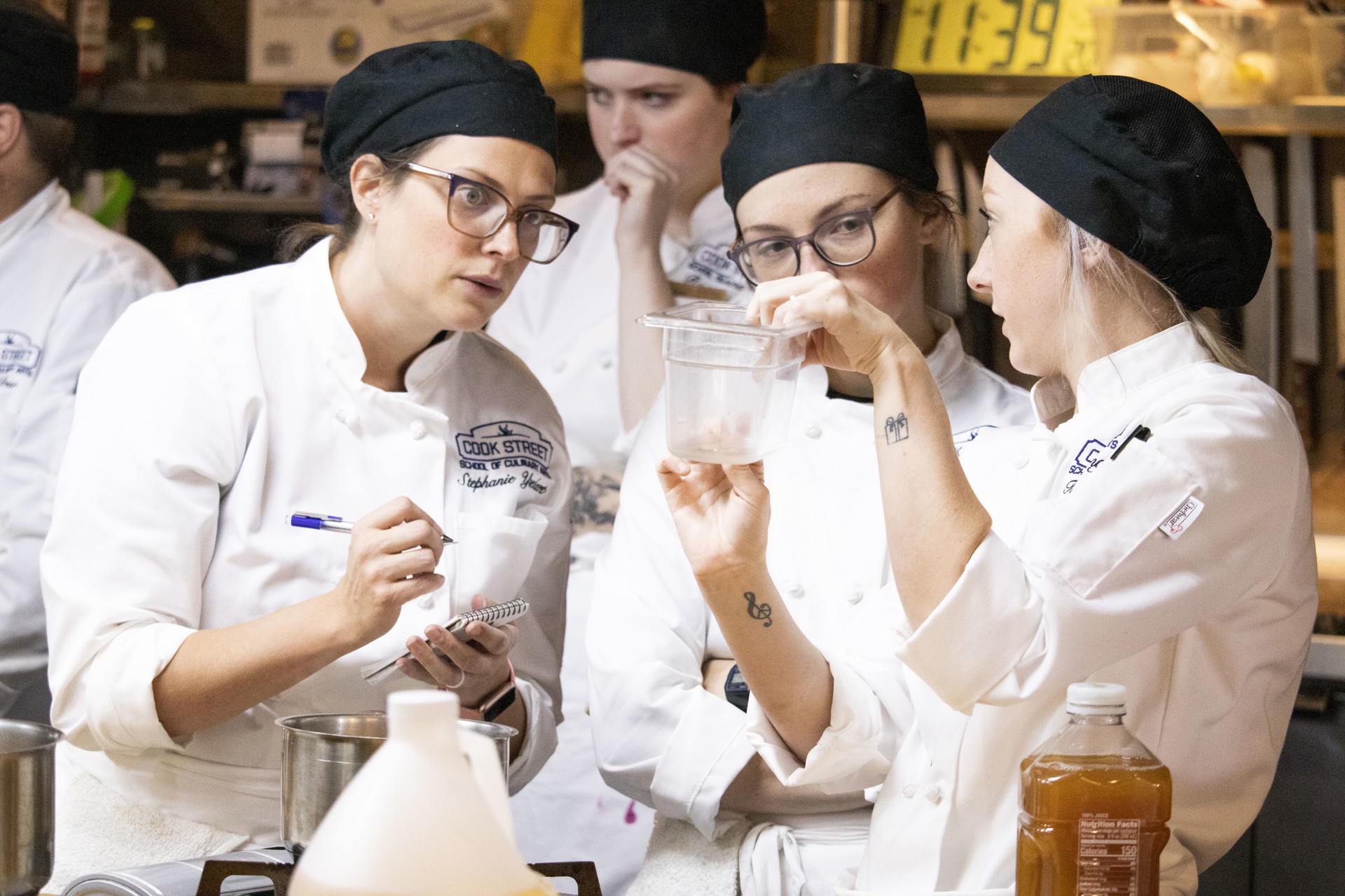 Three chefs in white jackets and black caps examine a clear container during a cooking class.