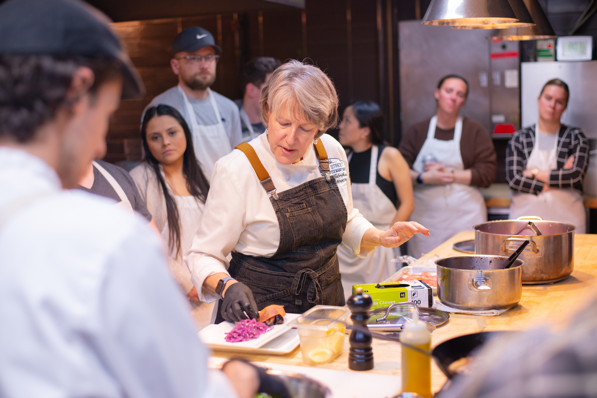 Chef in black apron demonstrating plating food to attentive cooking class students in kitchen.