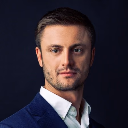 Portrait of a man with short brown hair and light beard wearing a white shirt and dark blue suit jacket against a dark background.