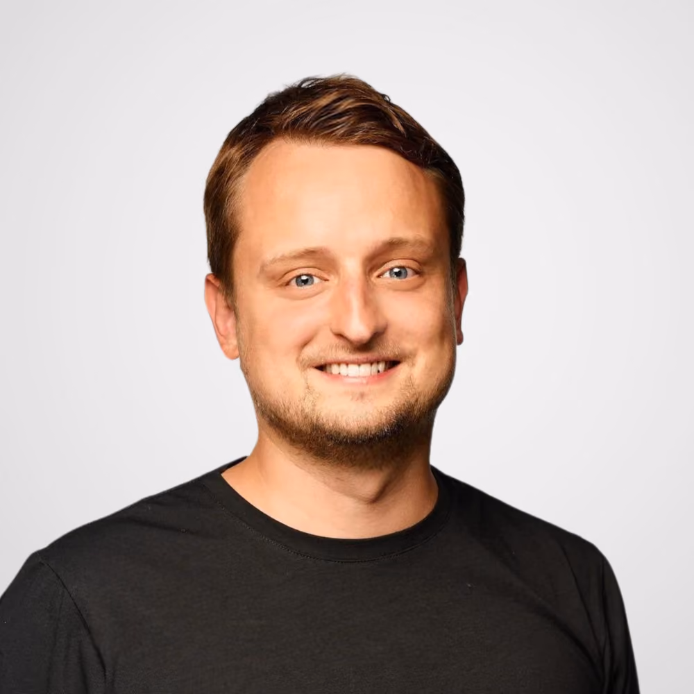 Smiling man with short brown hair and blue eyes wearing a black t-shirt against a light gray background.