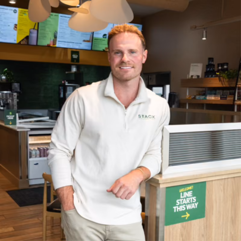 A man with short red hair and a light beard wearing a beige long-sleeve shirt with the word STACK on it stands inside a modern cafe near a counter with a sign that reads 'Welcome! Line starts this way' with an arrow.