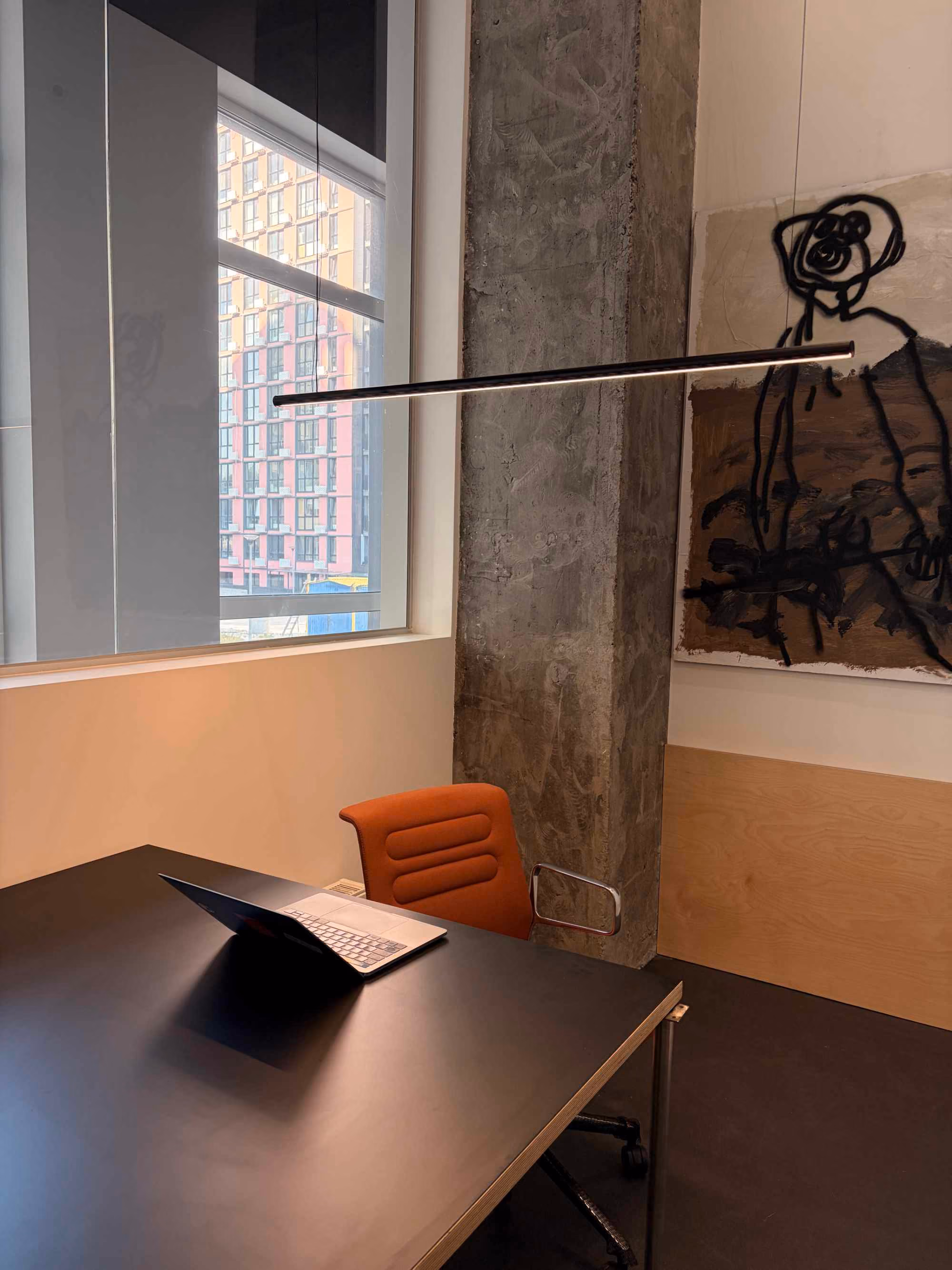 Minimalist office corner with a black desk holding an open laptop, an orange cushioned chair, a concrete pillar, a window showing an exterior building, and abstract wall art in black and brown tones.