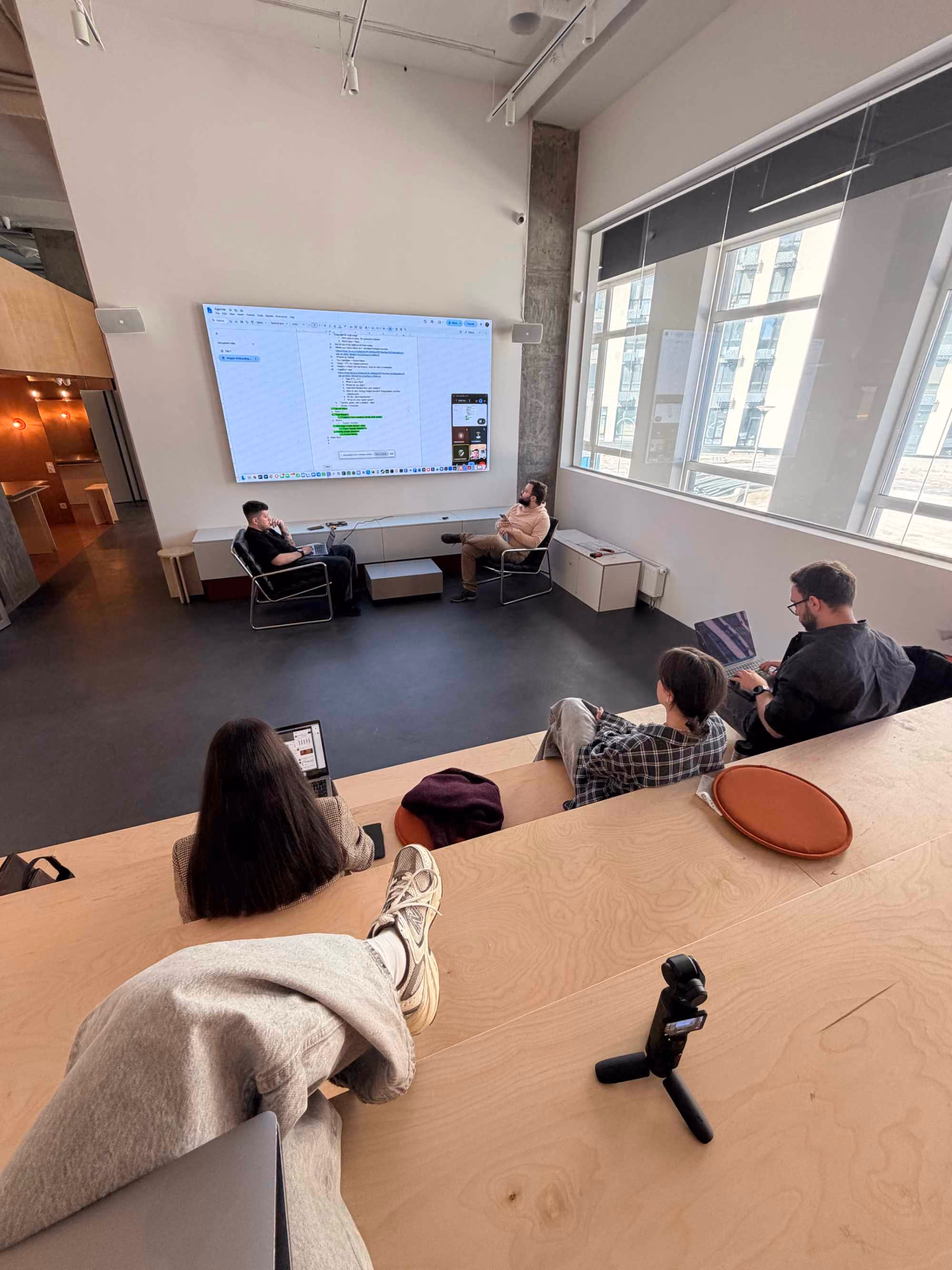 Modern conference room with five people: two men seated on chairs near a large wall-mounted screen displaying a digital document, and three people sitting on wooden tiered seating, two using laptops. Large windows on one side illuminate the room with natural light.