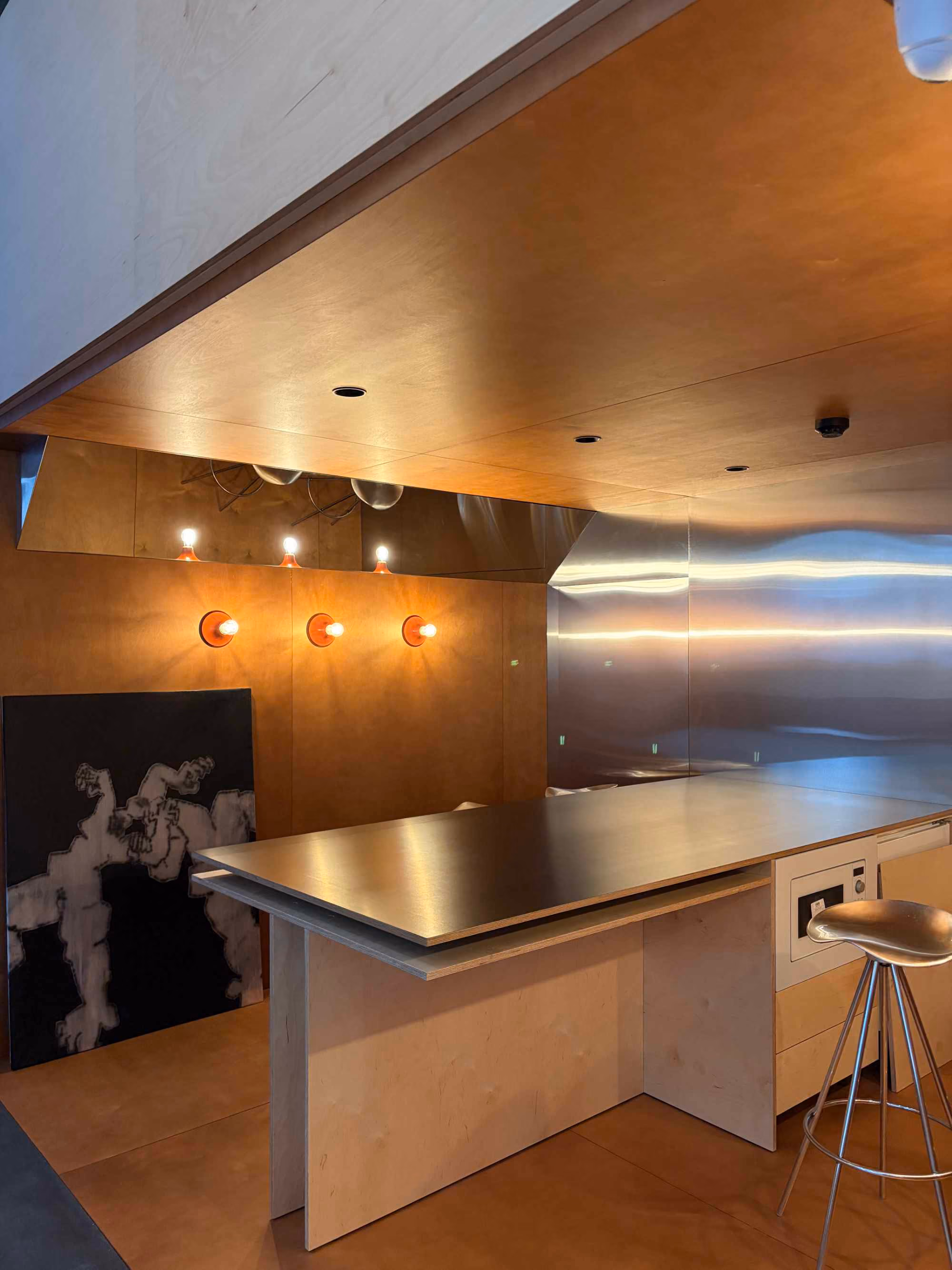 Modern kitchen area with wooden walls and ceiling, a large black countertop island, a metallic stool, three exposed light bulbs on the wall, a shiny metal backsplash, and black abstract artwork leaning against the wall.
