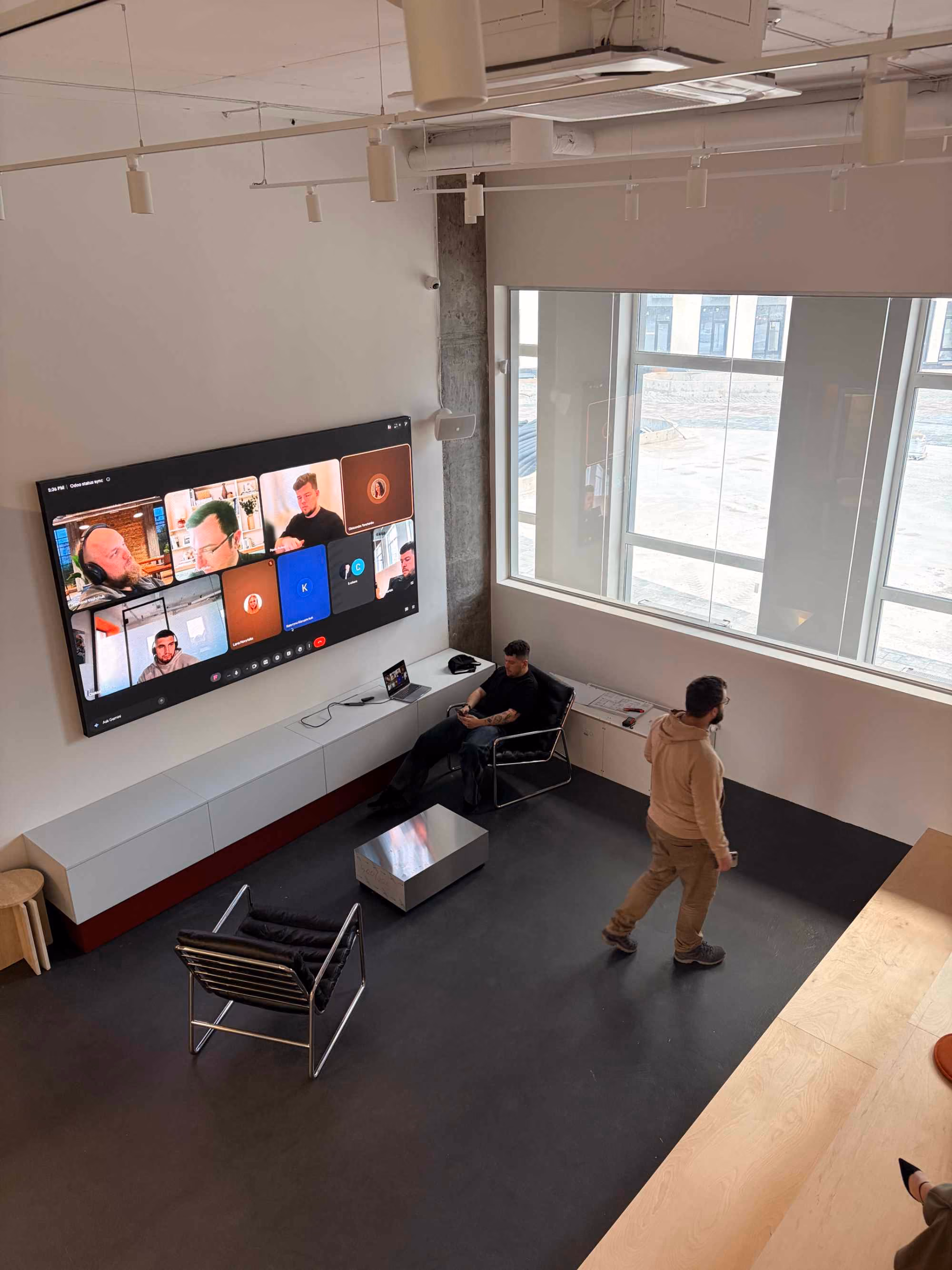 Modern office lounge with two men; one sitting in a black chair using a phone near a laptop, the other walking across the room, with a large screen on the wall displaying a virtual meeting with multiple participants.