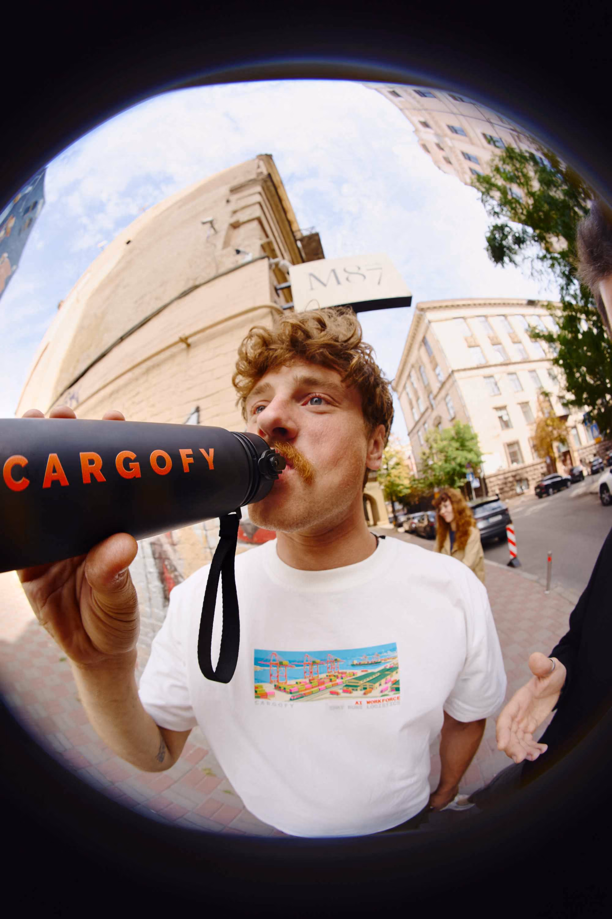 A man with curly hair and a mustache drinks from a black water bottle with the word CARGOFY in orange letters, standing on a city sidewalk with buildings and a woman in the background.