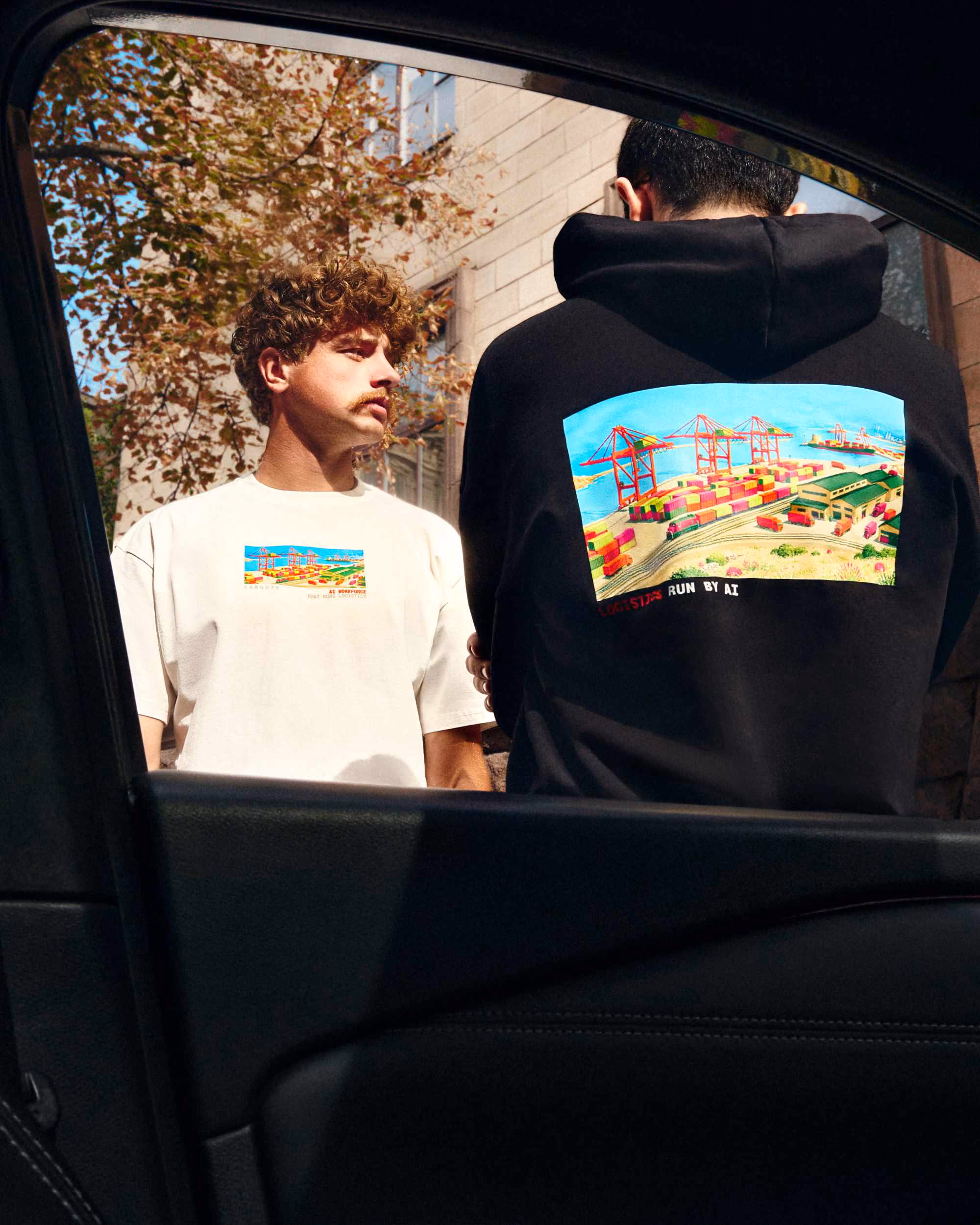 Two men standing outside near a building with fall foliage; one faces forward wearing a white t-shirt with a colorful graphic of a port with shipping containers and cranes, the other faces away wearing a black hoodie with the same graphic larger on the back.