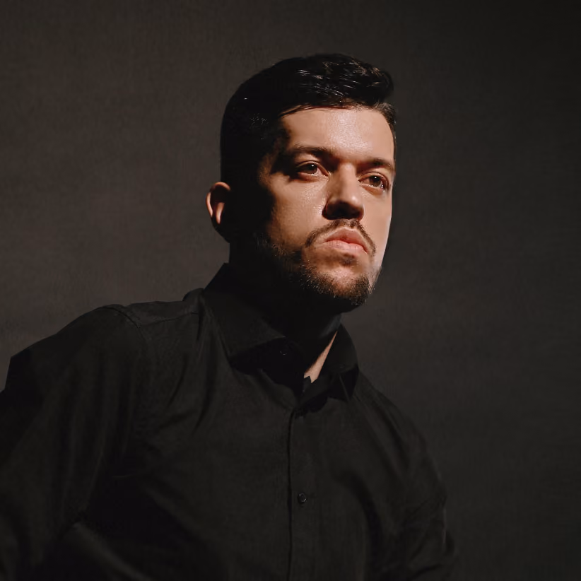 Portrait of a man with short dark hair and a trimmed beard wearing a black shirt, looking thoughtfully to the side against a dark background.
