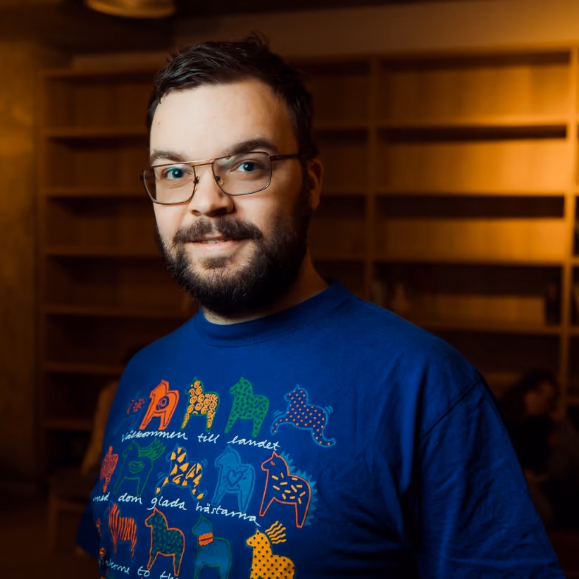 Man with short dark hair, beard, and glasses wearing a blue T-shirt with colorful horse designs and text, standing indoors with blurred wooden shelves in the background.